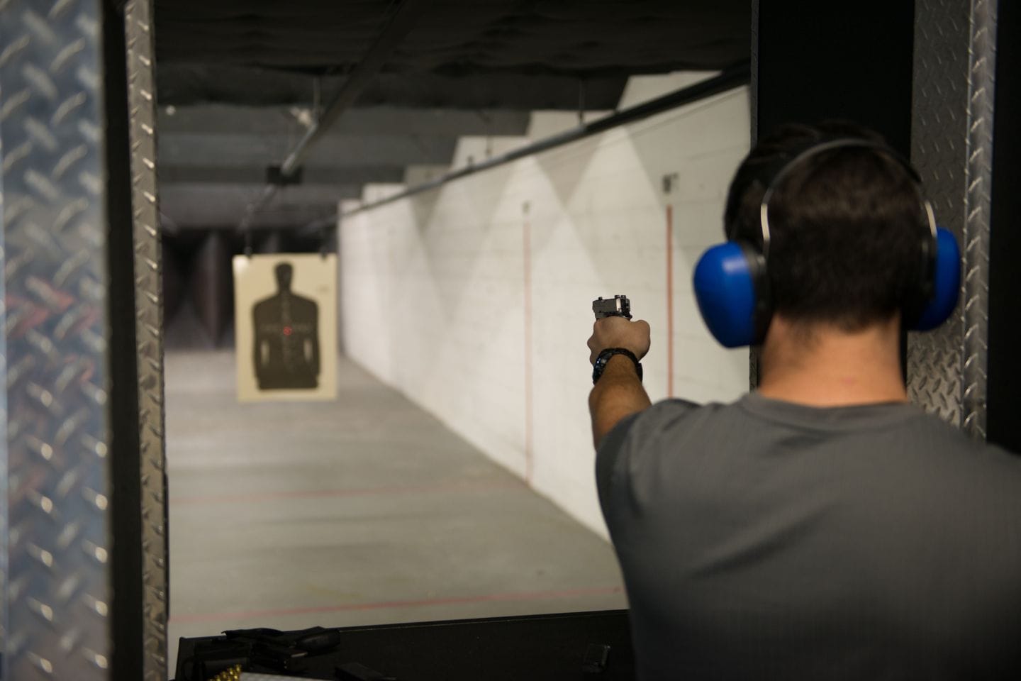 At an indoor shooting range, a person participates in active threat training, aiming a handgun at a paper target while wearing blue earmuffs.