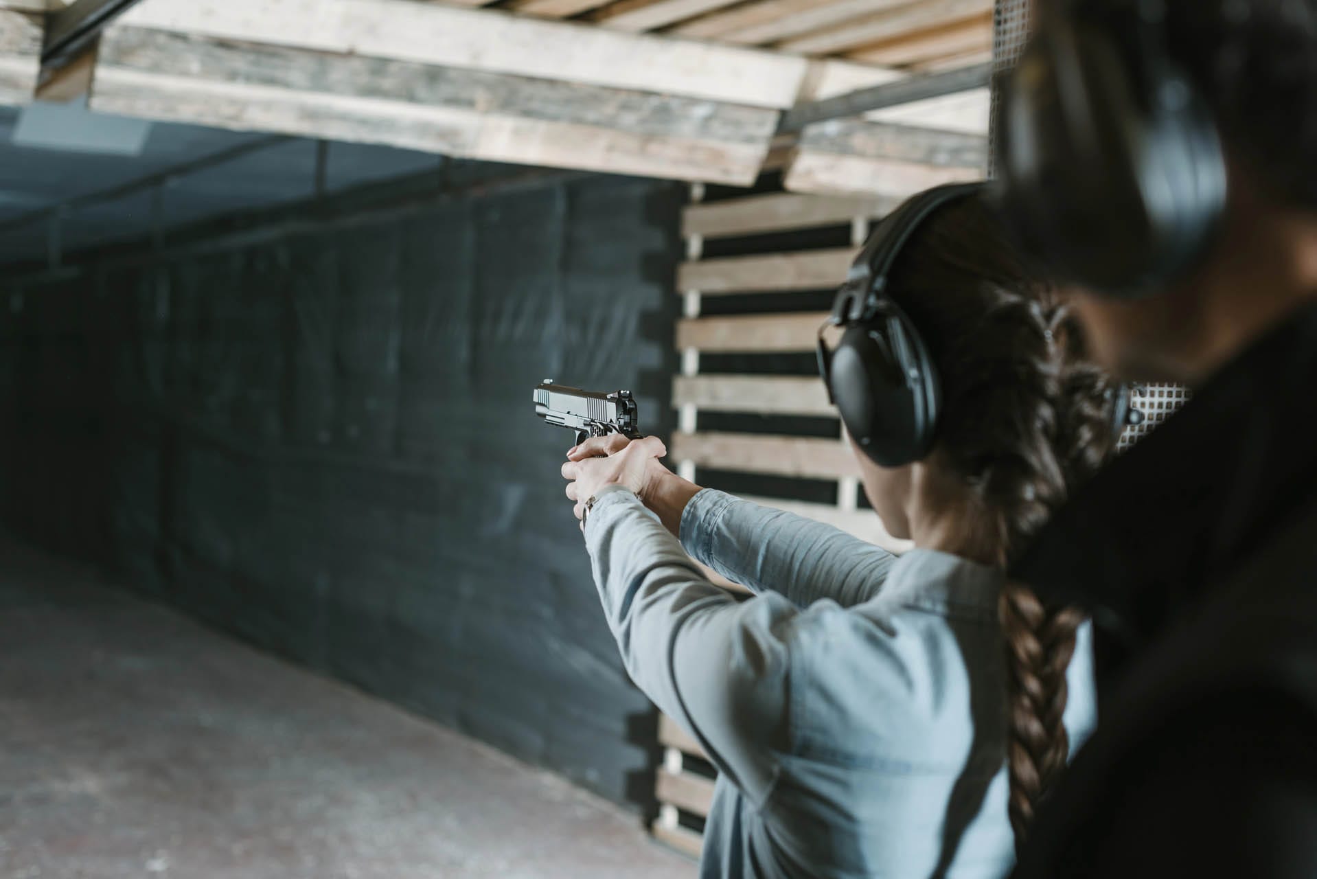 At the shooting range, a person focuses on active threat training, aiming a handgun as another observes from the side. Both wear protective ear muffs.