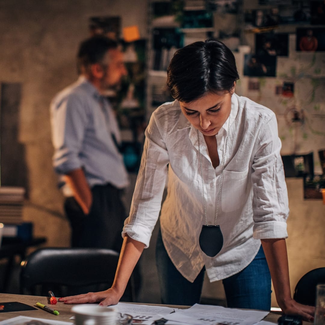 A person in a white shirt leans over a desk, examining documents, as part of providing investigation services. A blurred figure stands in the background near a wall covered with photos and papers.