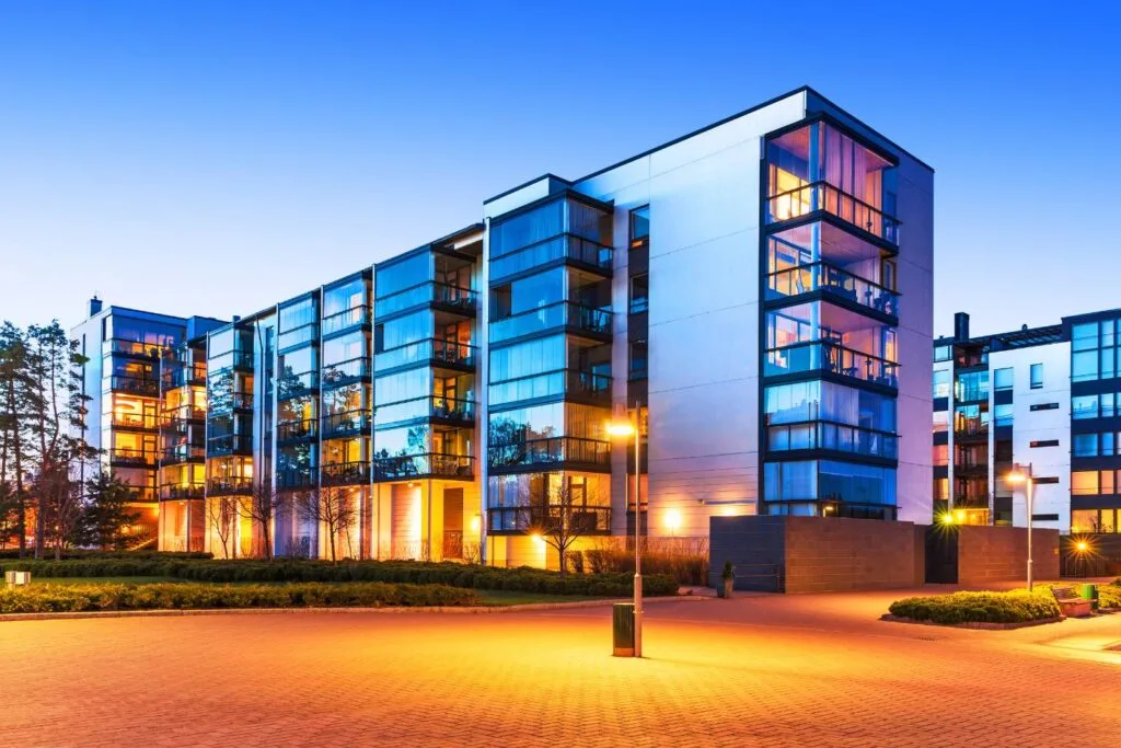 Modern apartment building with reflective glass windows, surrounded by landscaped greenery serving various industries, with warm exterior lighting at dusk.