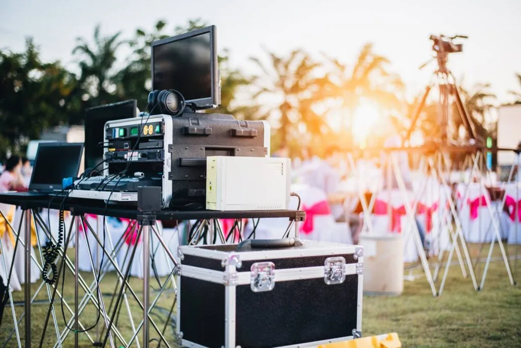 Outdoor event setup with audio-visual equipment, including monitors and cameras, on metal stands and tables. The scene reflects the diverse industries served as sunlight filters through trees in the background.