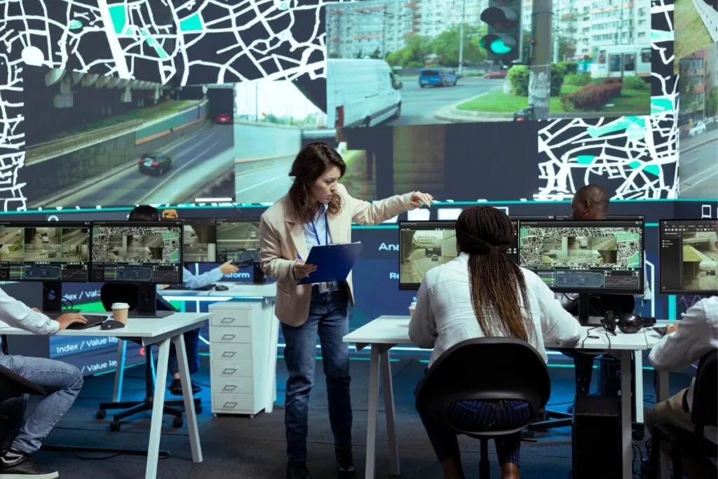 Identifying Issues With Your Current GPS Tracking System 1 A woman with a clipboard instructs three people seated at computer monitors in a control room displaying traffic footage and maps on large screens, seamlessly integrating data from the GPS tracking system.
