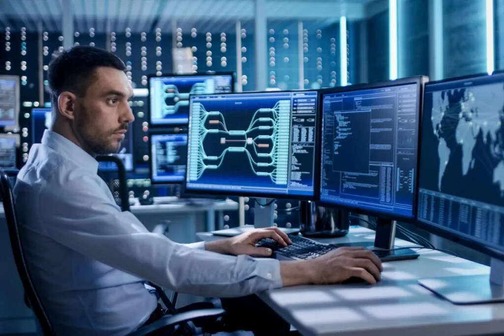Identifying Issues With Your Current GPS Tracking System 5 A man in a white shirt sits at a desk with multiple computer screens displaying complex graphics, code, and GPS tracking system data in a modern office setting.