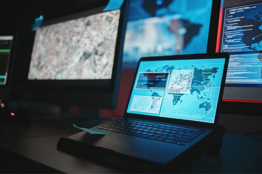 Identifying Issues With Your Current GPS Tracking System 10 The laptop, featuring a GPS tracking system, displays maps and data, surrounded by multiple monitors showcasing various technical screens in a dimly lit room.