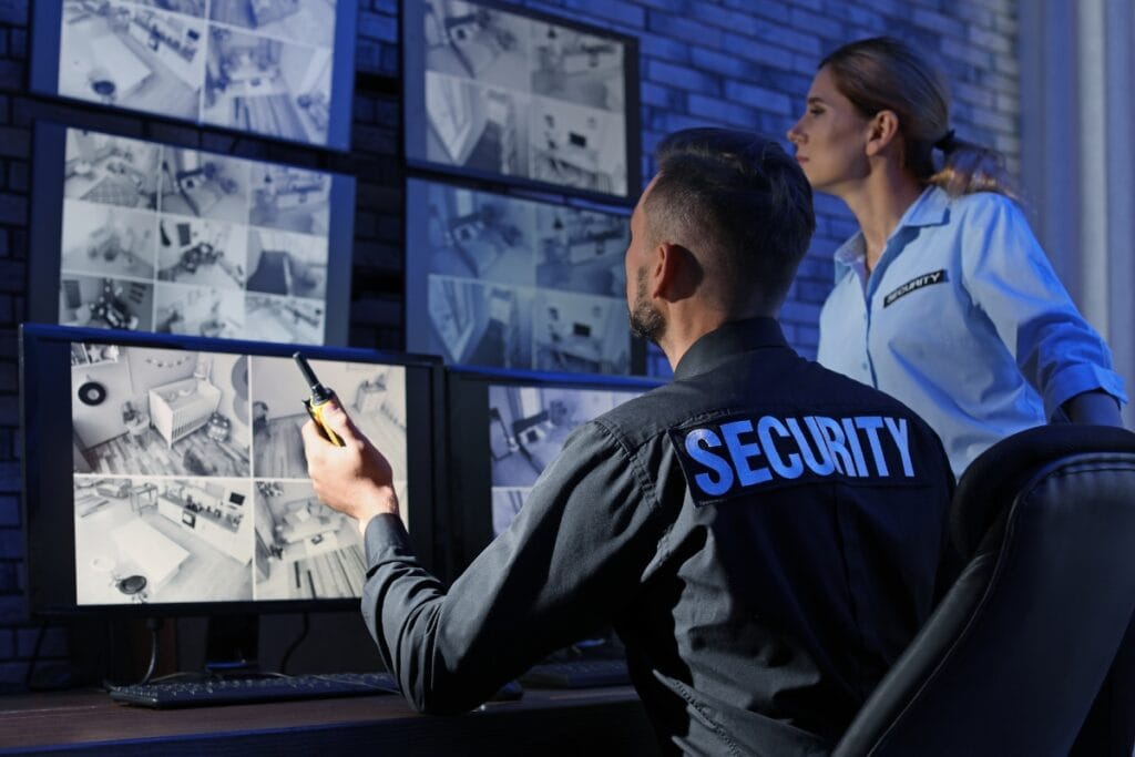 In a professional security service control room, two personnel closely monitor multiple CCTV screens. The man, holding a walkie-talkie, coordinates efficiently while the woman intently observes the displays, ensuring utmost vigilance and safety at all times.