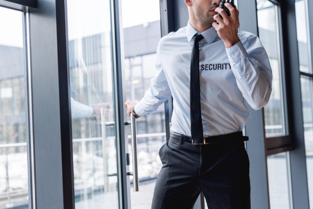 A professional security service guard in uniform speaks into a walkie-talkie while standing near a glass door in a modern building.