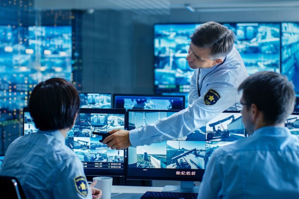 Three security personnel in a control room, representing a professional security service, are monitoring multiple screens displaying surveillance footage. One person is pointing at a screen while discussing strategies with two colleagues.