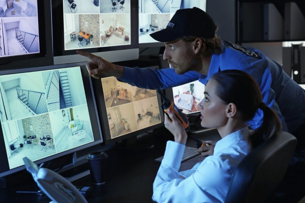 Two security personnel from the professional security service monitor multiple CCTV feeds on large screens. One is pointing, and the other is holding a walkie-talkie.