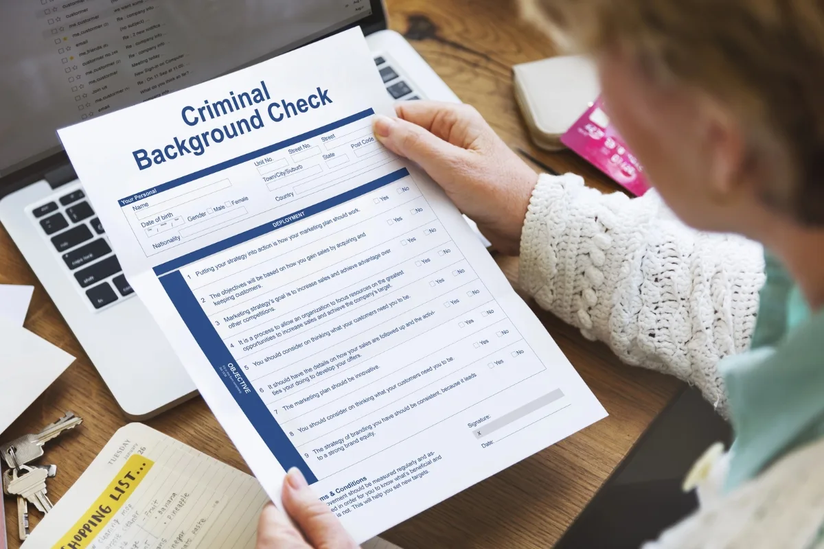 A person holds a printed criminal background check form over a laptop, with keys and a notepad nearby, highlighting the meticulous hiring process employers undertake to ensure safety and reliability in their workforce.