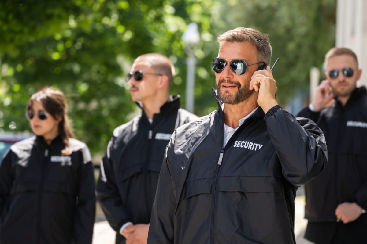 Four armed and unarmed security guards in black jackets and sunglasses, with one speaking into a walkie-talkie, stand outdoors on a sunny day.