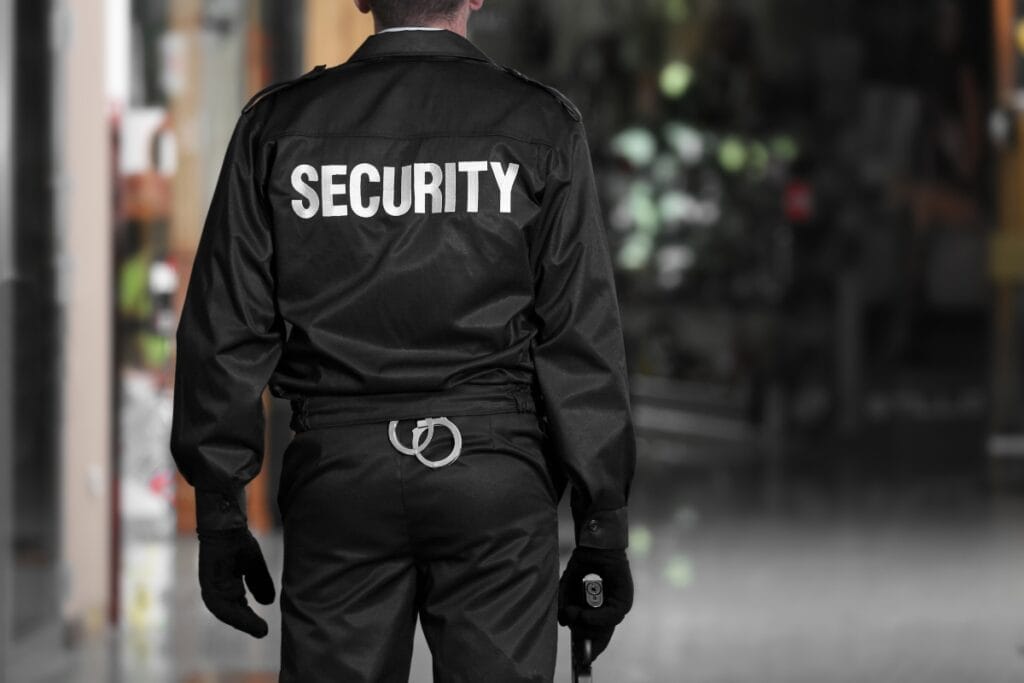 A security guard stands indoors, facing away from the camera. The word "SECURITY" is printed in large white letters on the back of the uniform, highlighting their role among armed and unarmed security guards. The guard holds a flashlight.