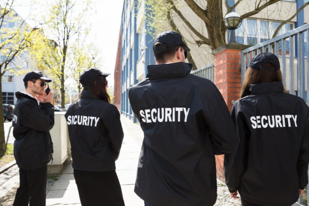 Four Armed and Unarmed Security Guards in black uniforms stand outside near a fence and brick pillar, their backs to the camera.