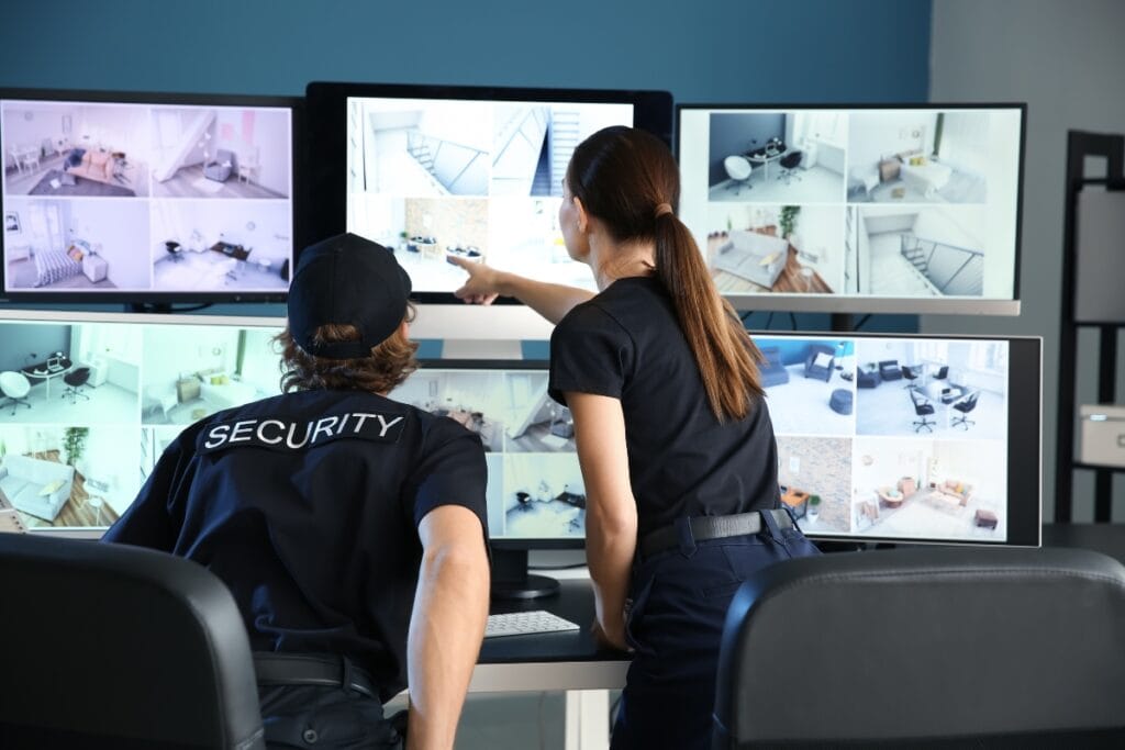 Two security officers, specializing as armed and unarmed security guards, monitor multiple surveillance camera feeds displayed on several large screens in a control room.