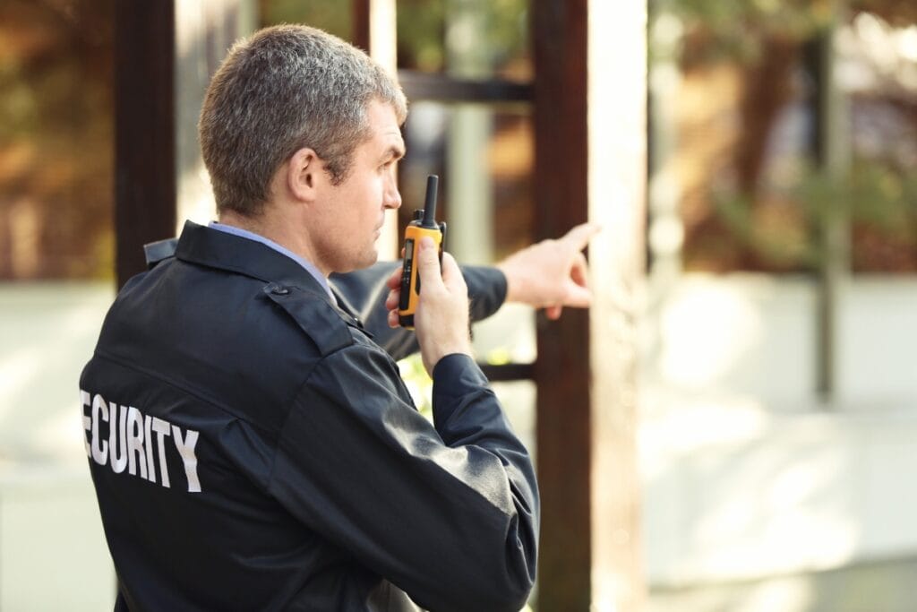 An armed and unarmed security guard holding a walkie-talkie stands outdoors, looking and pointing to the right.