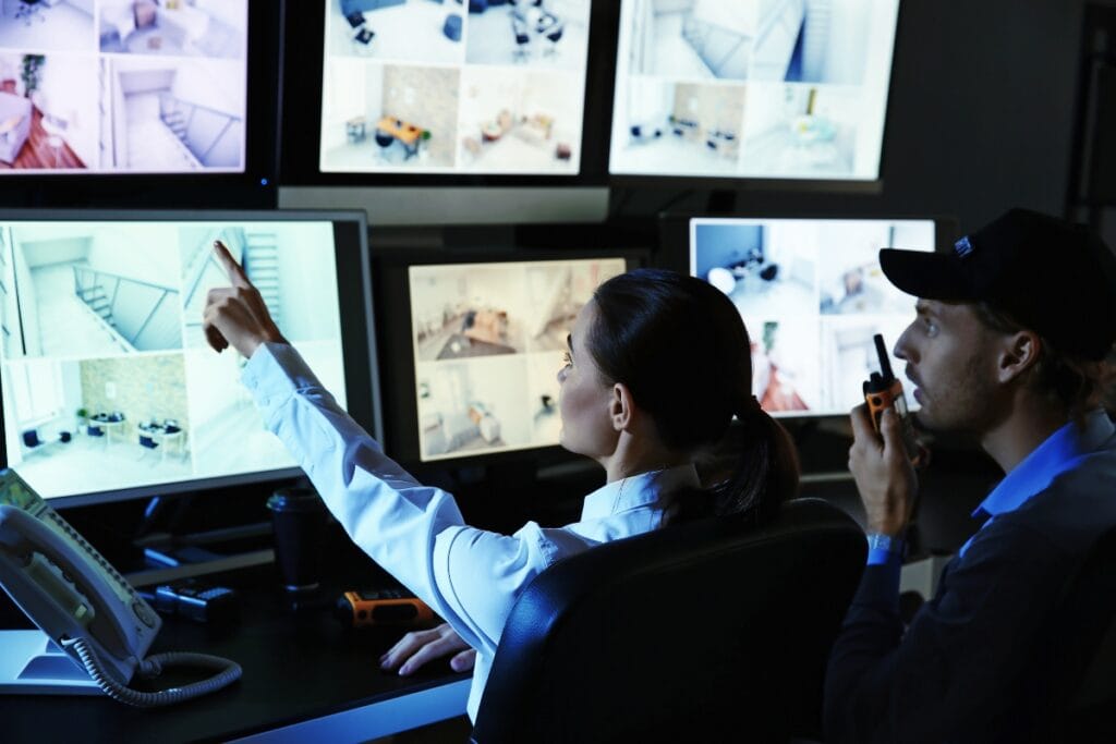 Two security personnel monitor multiple surveillance camera feeds on screens; one points at a screen while the other speaks into a walkie-talkie, ensuring top-notch surveillance services.