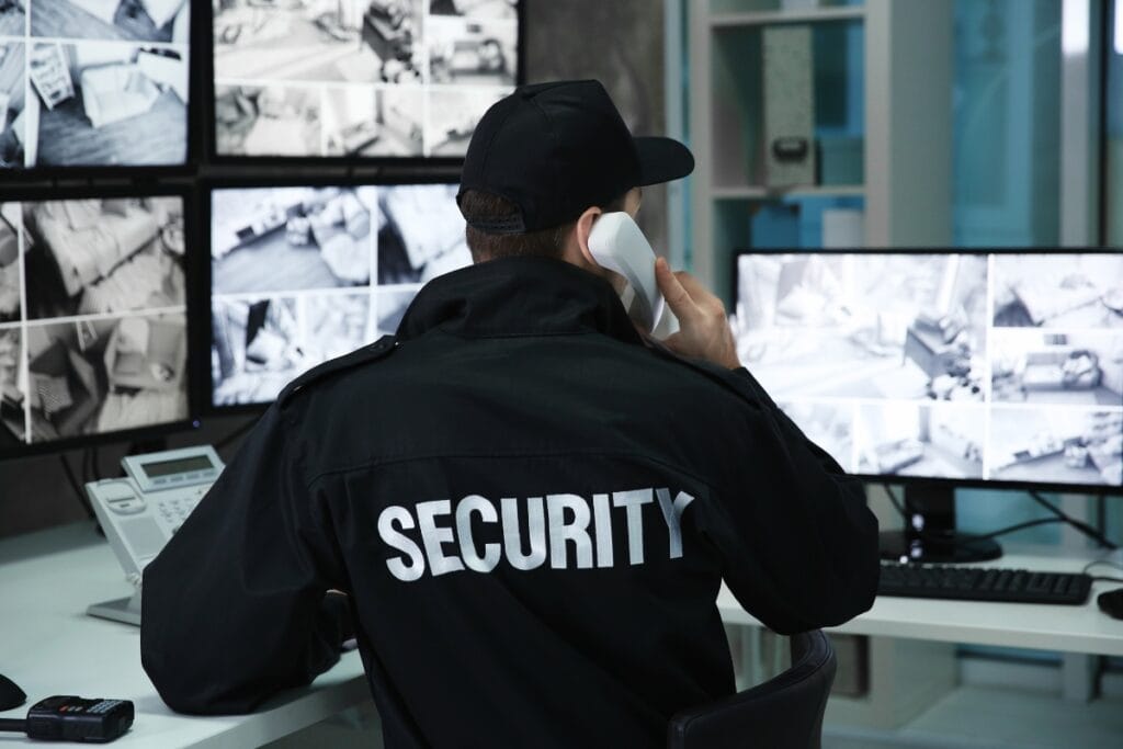 A security guard in uniform monitors multiple surveillance screens and speaks on a landline phone in a control room, ensuring top-notch surveillance services.