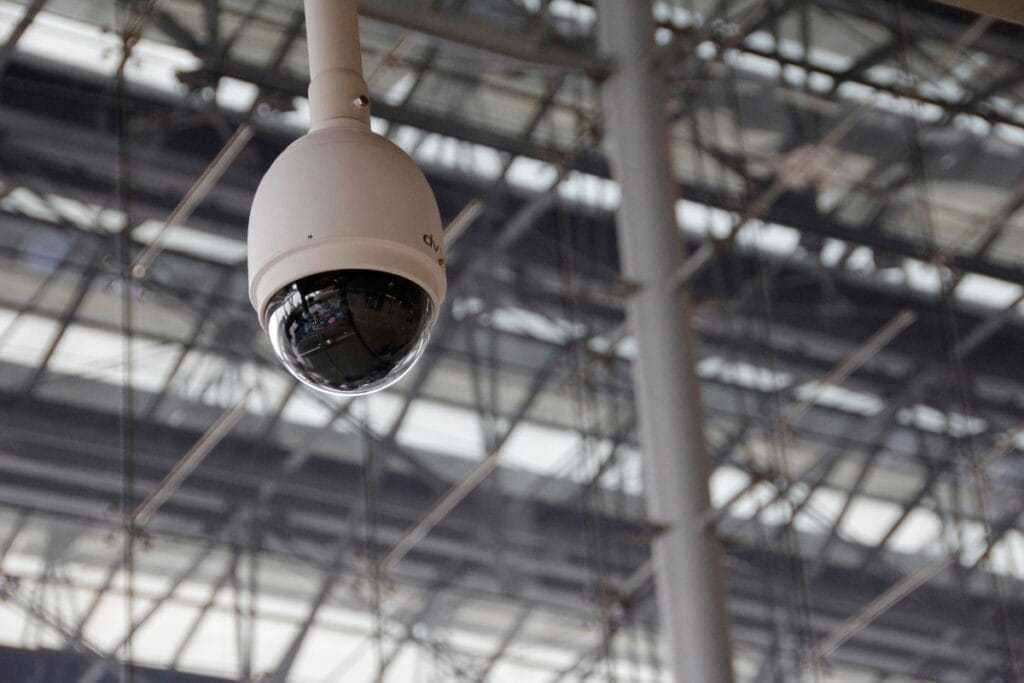 A dome security camera, part of comprehensive surveillance services, is mounted on the ceiling of a large, industrial-style building with metal beams and glass panels.