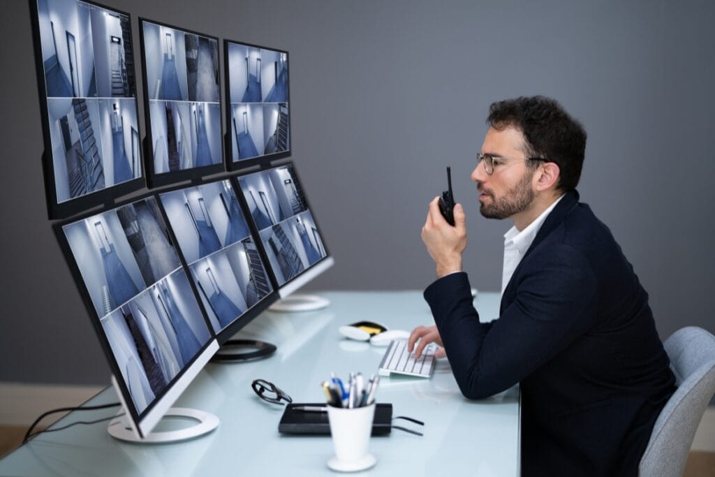A man in a suit monitors multiple security camera feeds on six screens at a desk, providing surveillance services while holding a walkie-talkie and typing on a keyboard.