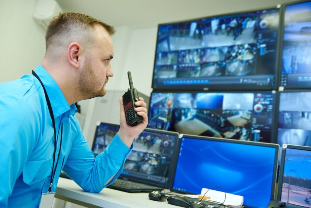 Man in blue shirt using two-way radio monitors security camera footage in a control room, showcasing professional surveillance services.