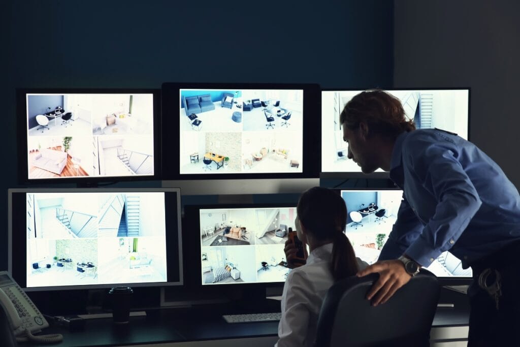 Two people monitor multiple security camera feeds on large computer screens in a dimly lit control room, showcasing professional surveillance services.