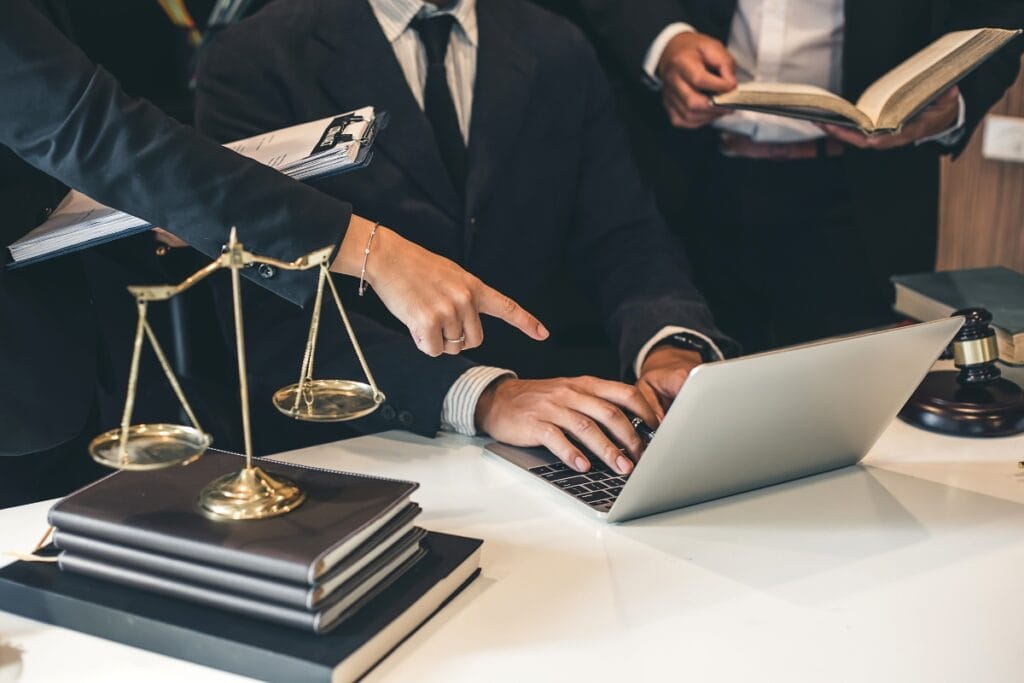 Three people in business attire work at a desk with a laptop, legal books, scales of justice, and a gavel; one person points at the screen as they discuss Litigation Support Investigations.