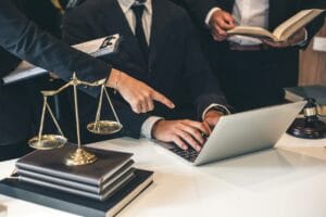 Blog 2 Three people in business attire work at a desk with a laptop, legal books, scales of justice, and a gavel; one person points at the screen as they discuss Litigation Support Investigations.