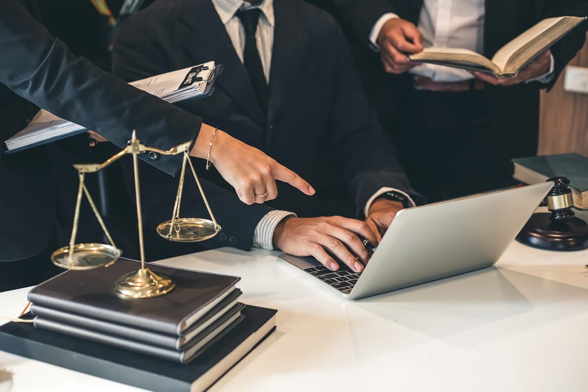 Three people in business attire work at a desk with a laptop, legal books, scales of justice, and a gavel; one person points at the screen as they discuss Litigation Support Investigations.