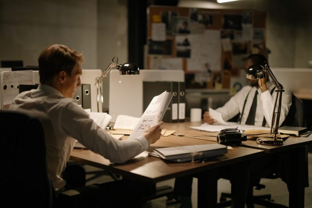 Litigation Support Investigations: How Private Investigators Assist Attorneys 3 Two men wearing shirts and ties work at desks in a dimly lit office, examining documents under desk lamps with files, papers, and a bulletin board in the background—immersed in litigation support investigations.