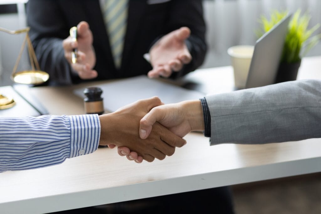 Litigation Support Investigations: How Private Investigators Assist Attorneys 8 Two people shake hands across a desk, with a person in a suit seated in the background, legal documents, a gavel, and scales of justice visible—an ideal setting for litigation support investigations.