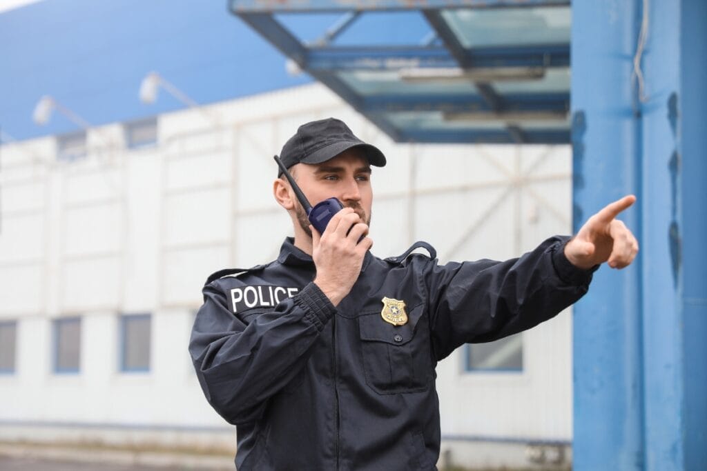 Mobile Security Patrol: How Mobile Security Can Protect Your Business 1 A police officer in uniform, part of a mobile security patrol, talks into a radio and points with his right hand outside an industrial building.