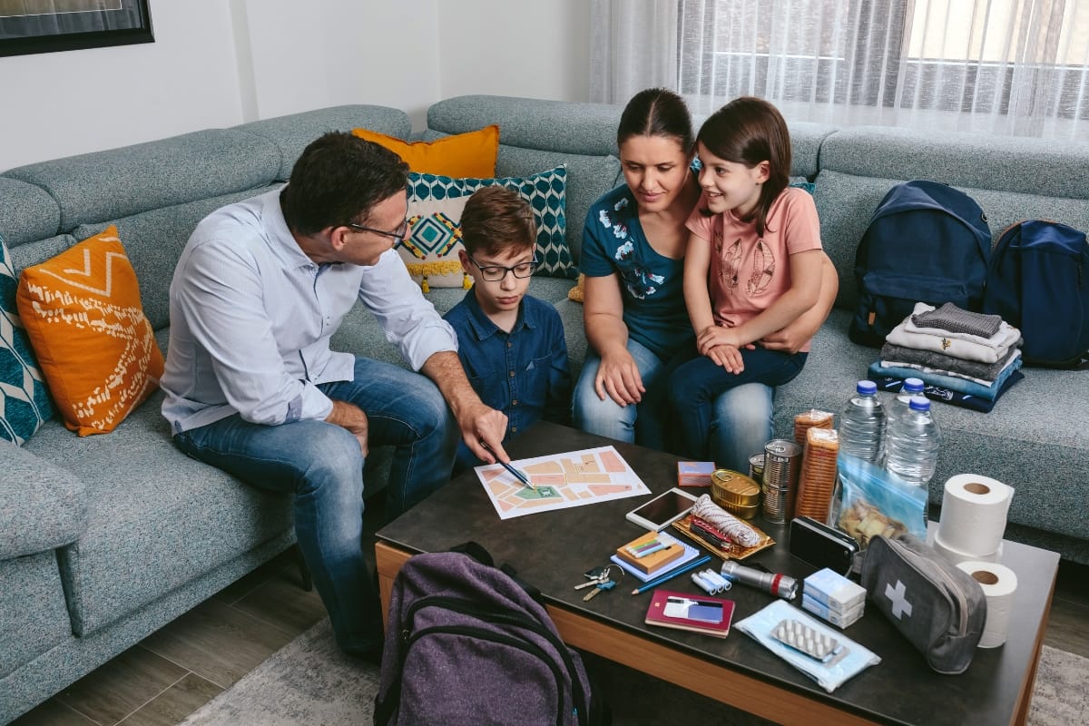 A family of four sits in a living room, discussing an evacuation plan with emergency supplies and bottled water on the table, practicing active threat training together.