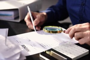 Blog 9 Person examining an invoice with a magnifying glass while writing, with a calculator and paperwork on the desk—an activity often seen during corporate investigations.