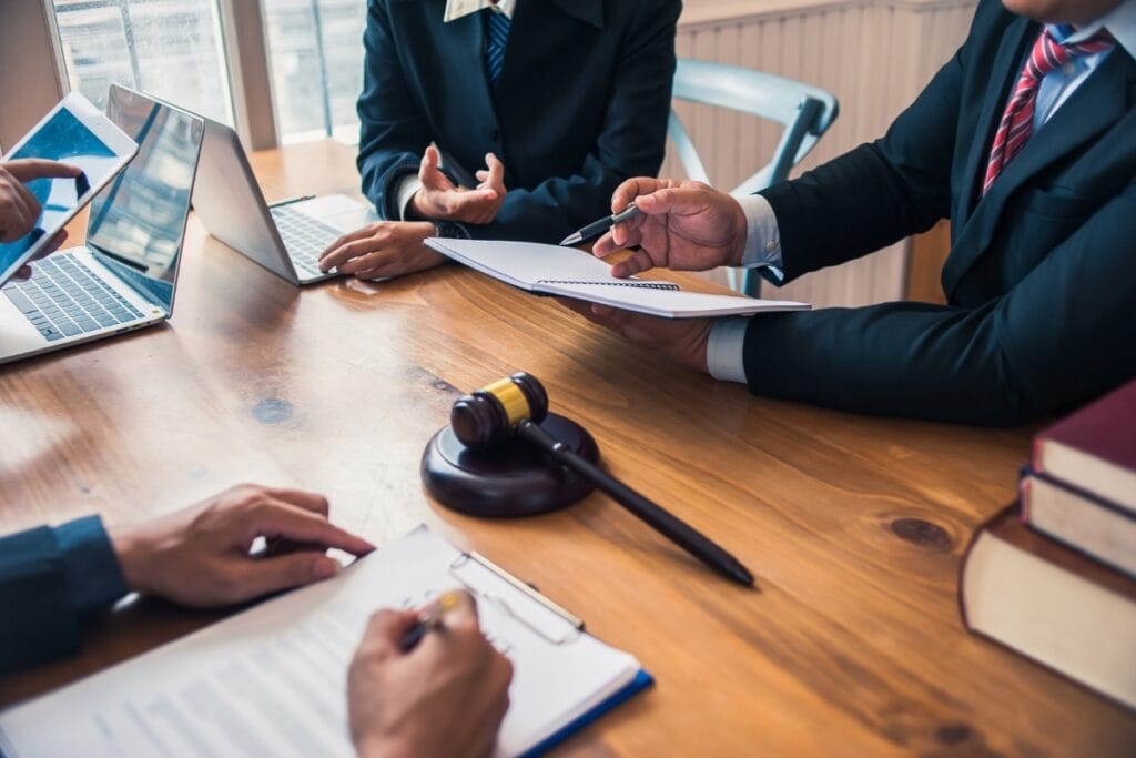 Corporate Investigations: How Businesses Can Detect Fraud and Theft 5 Three people in business attire sit at a table with laptops, documents, and a judge’s gavel, discussing paperwork related to corporate investigations in a legal or professional setting.