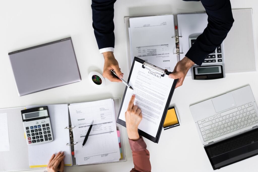 Corporate Investigations: How Businesses Can Detect Fraud and Theft 6 Two people exchanging a clipboard with a contract over a desk covered with binders, documents, calculators, and a laptop—an environment typical for in-depth corporate investigations.