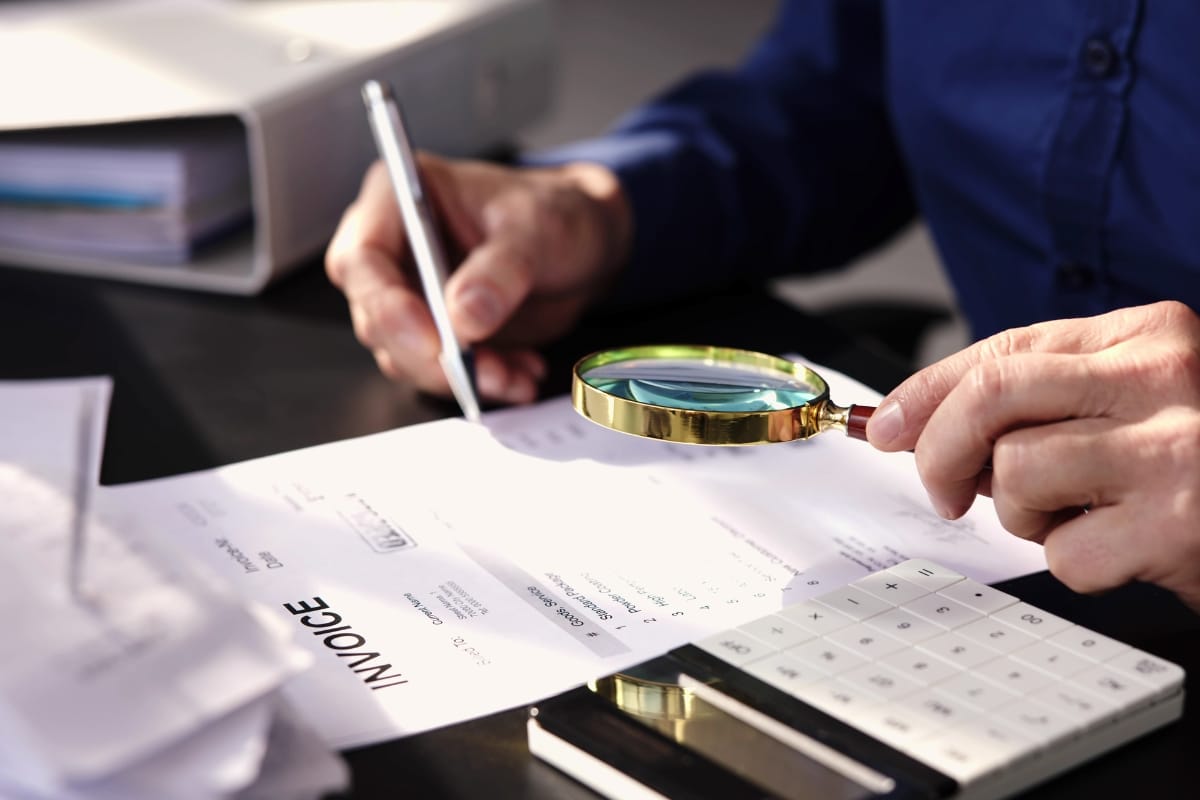 Person examining an invoice with a magnifying glass while writing, with a calculator and paperwork on the desk—an activity often seen during corporate investigations.