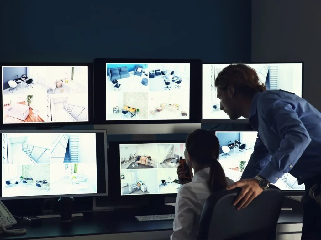 Two people in a control room monitor multiple security camera feeds on six large screens, carefully analyzing footage for infidelity investigations.