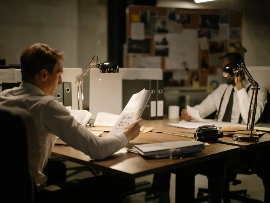 Two men in dress shirts sit at desks in a dimly lit office, examining documents related to infidelity investigations under desk lamps, with a bulletin board visible in the background.