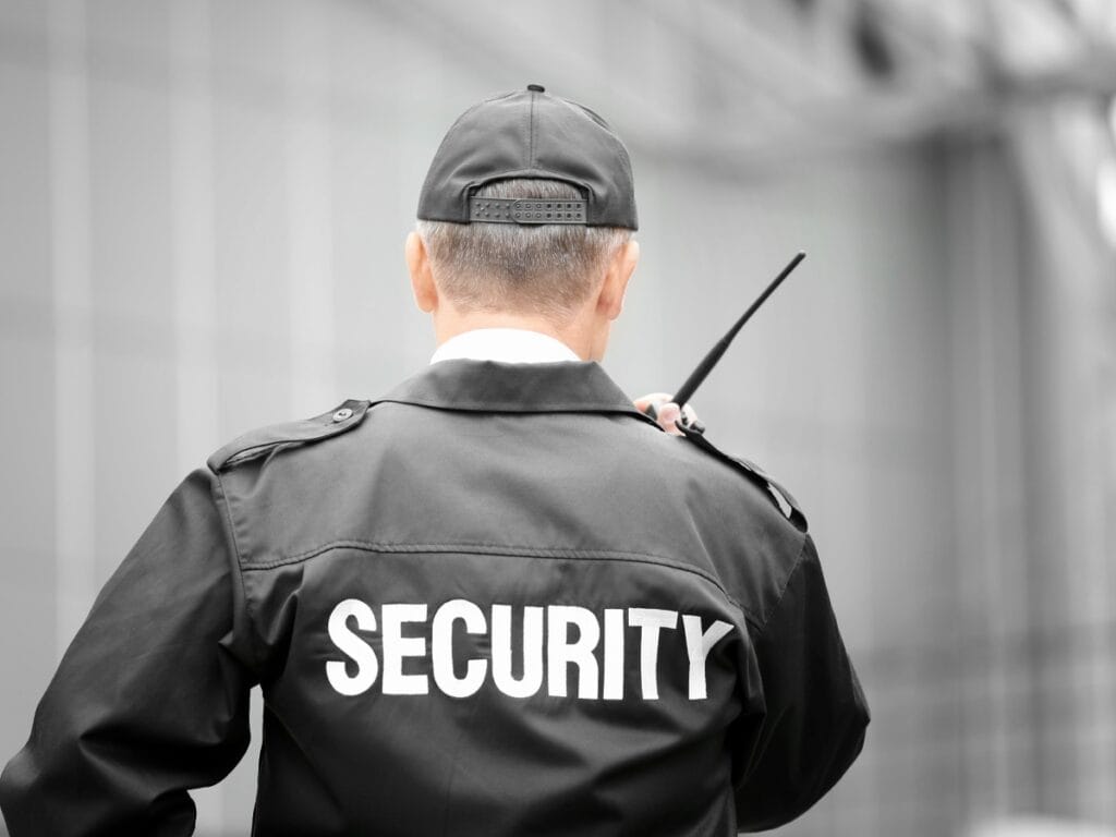 Security Guards in Georgia: Why Demand is Increasing for Business Protection 8 A security guard wearing a uniform and cap is seen from behind, holding a walkie-talkie in front of a modern building—typical of Security Guards in Georgia ensuring safety and vigilance.