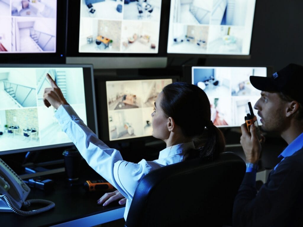 Security Guards in Georgia: Why Demand is Increasing for Business Protection 11 Two Security Guards in Georgia monitor multiple camera feeds on large screens; one points at a screen while the other holds a walkie-talkie.