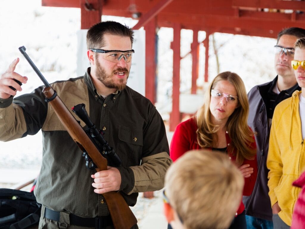 Atlanta Firearm Training: Essential Tips for Responsible Gun Owners 2 A man wearing safety glasses holds a rifle and gestures while speaking to a group of people, who listen attentively during an Atlanta Firearm Training session at an outdoor shooting range.