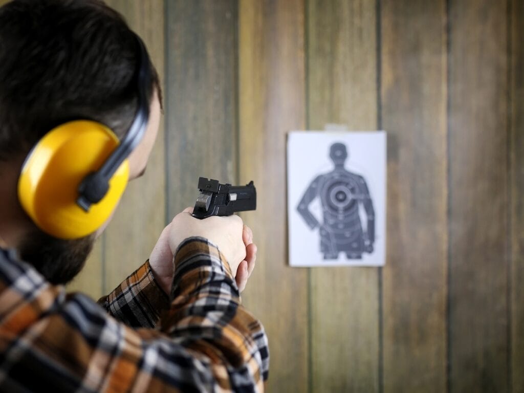Atlanta Firearm Training: Essential Tips for Responsible Gun Owners 4 A person wearing yellow ear protection aims a handgun at a paper target with a human silhouette on a wooden wall background during Atlanta Firearm Training.