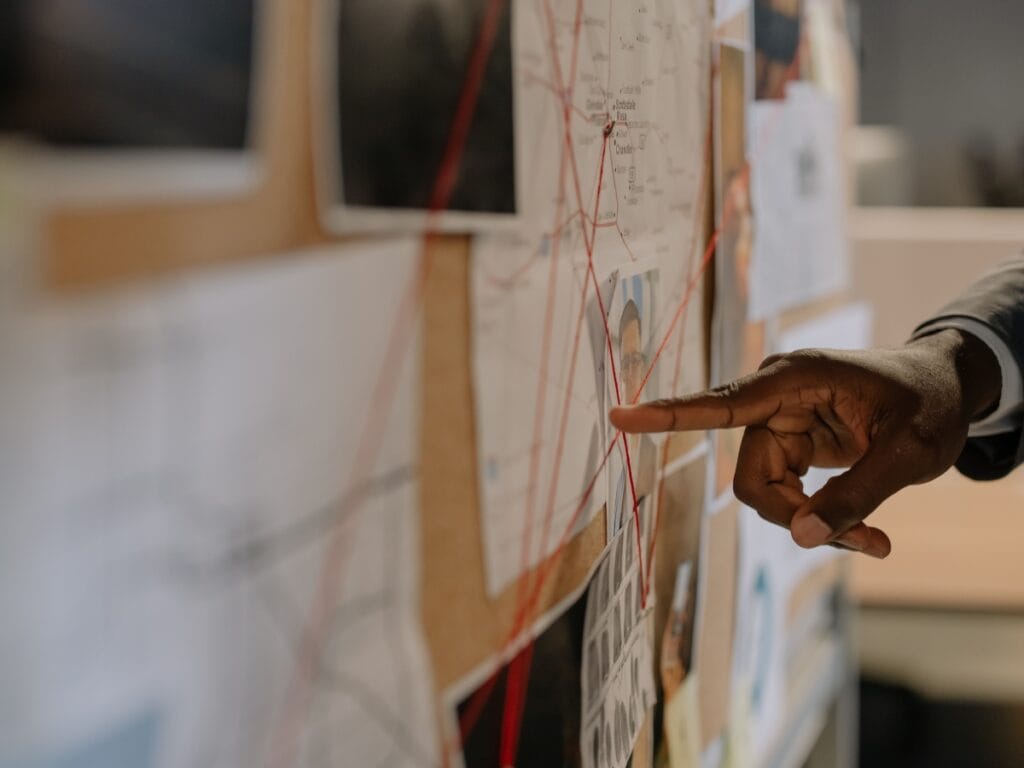 A hand points at a map on a corkboard covered with documents, photographs, and red string connecting various points, illustrating the detailed Private Investigation Process.