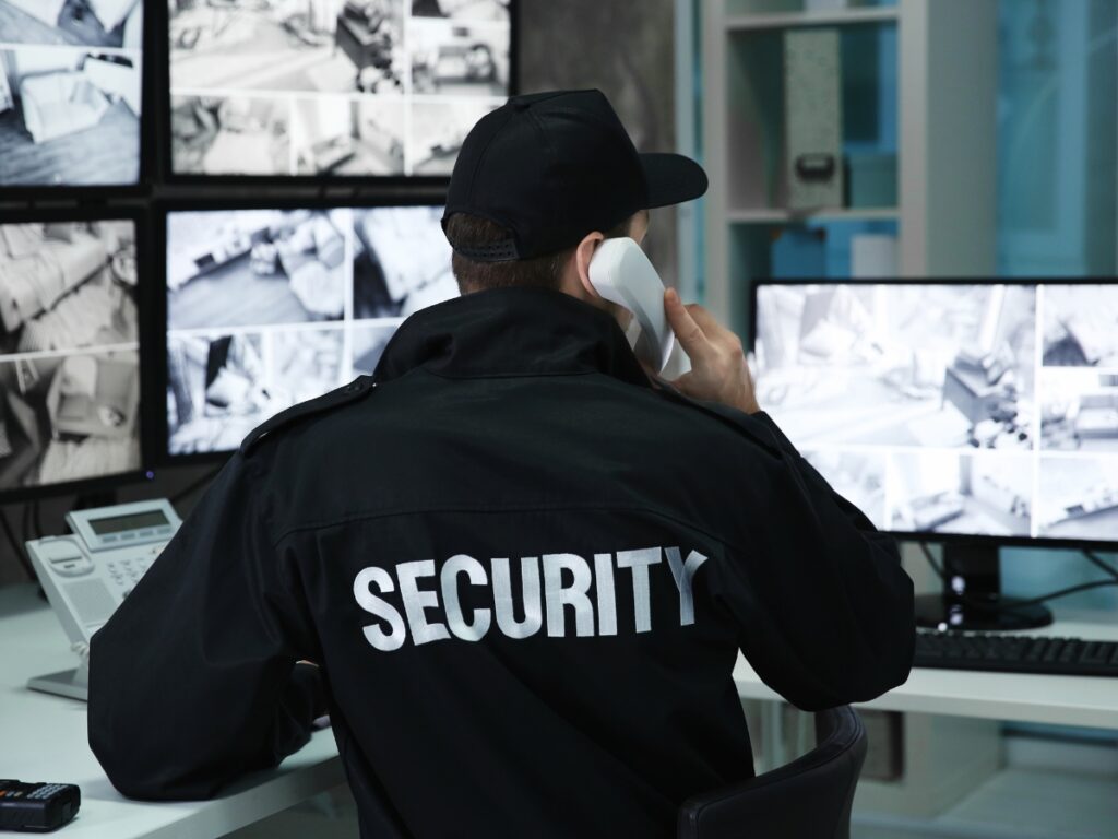 Holiday Security: How to Protect Your Store from Seasonal Theft 3 A security guard wearing a uniform and cap sits at a desk monitoring multiple surveillance screens while discussing holiday security measures on a landline phone.