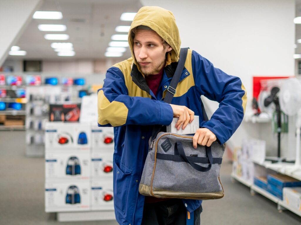 Holiday Security: How to Protect Your Store from Seasonal Theft 5 A man in a blue and tan hooded jacket appears to be putting an item into a gray bag inside a retail store, highlighting the importance of Holiday Security during busy shopping seasons.