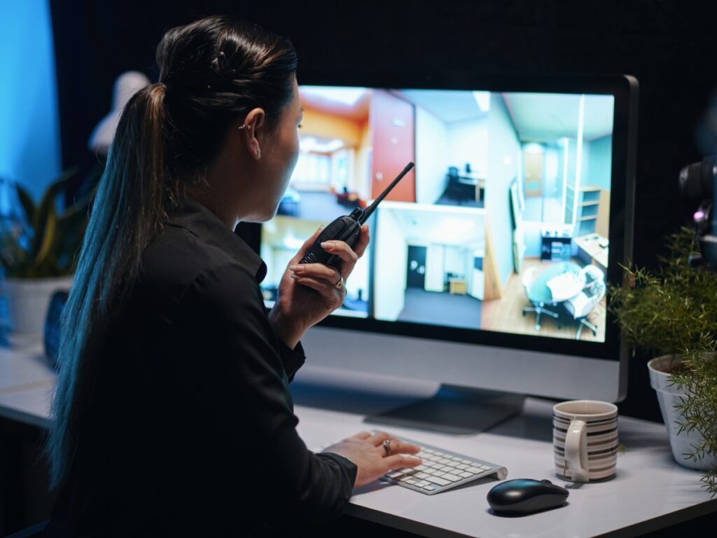A person holding a walkie-talkie monitors multiple security camera feeds on a computer screen at a desk, showcasing the vigilance and expertise of professional Security Consulting Services.