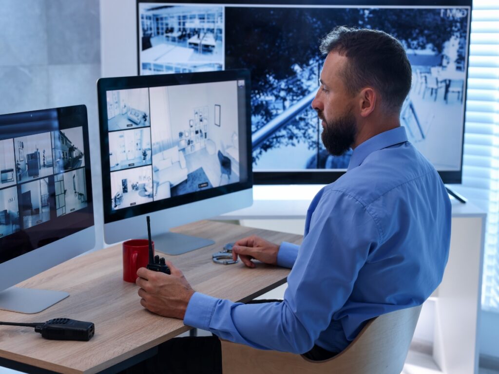 A man in a blue shirt sits at a desk monitoring multiple security camera feeds on computer screens, holding a walkie-talkie, exemplifying the vigilance provided by professional Security Consulting Services.