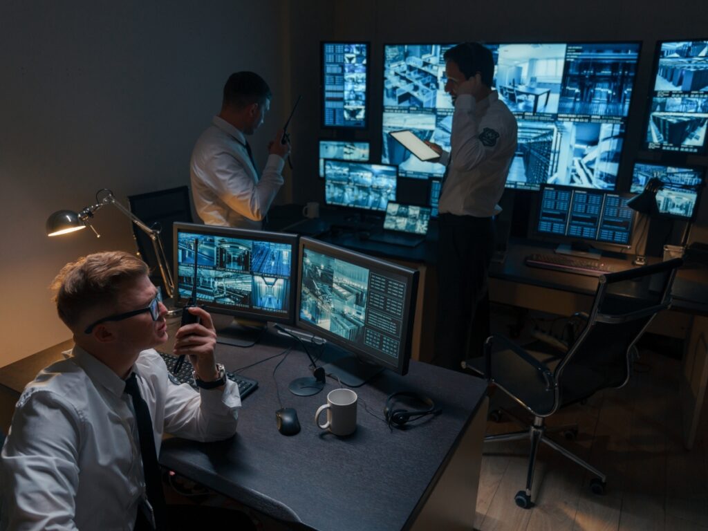 Three security personnel monitor multiple surveillance screens and communicate via radios in a dimly lit control room, showcasing the vigilance behind professional Security Consulting Services.