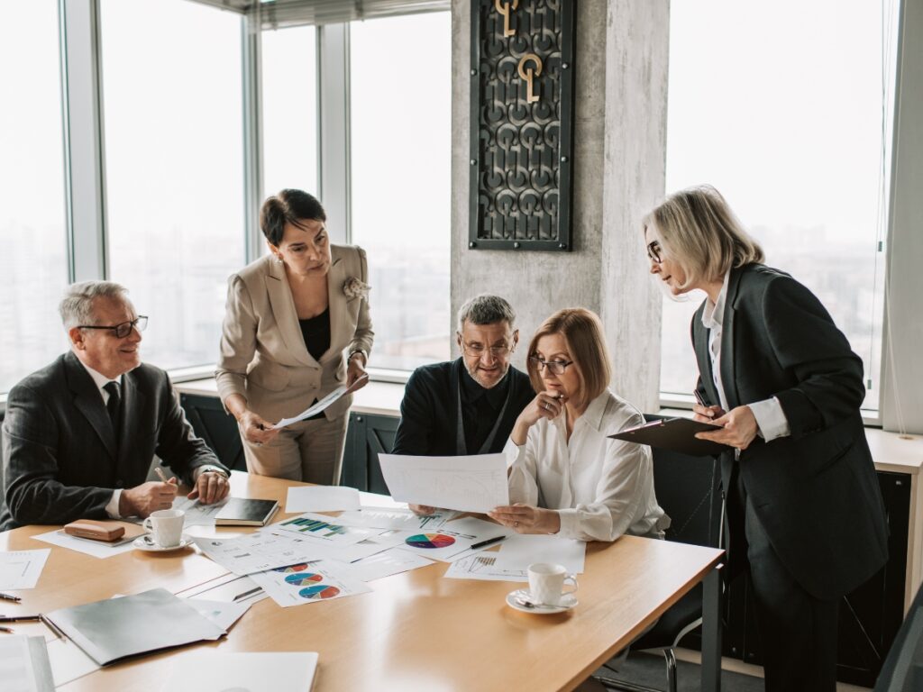 Business Fraud Investigations: How Private Investigators Help Protect Your Company 1 Five people in business attire are gathered around a conference table, reviewing documents and charts related to business fraud investigations during a meeting in a modern office setting.