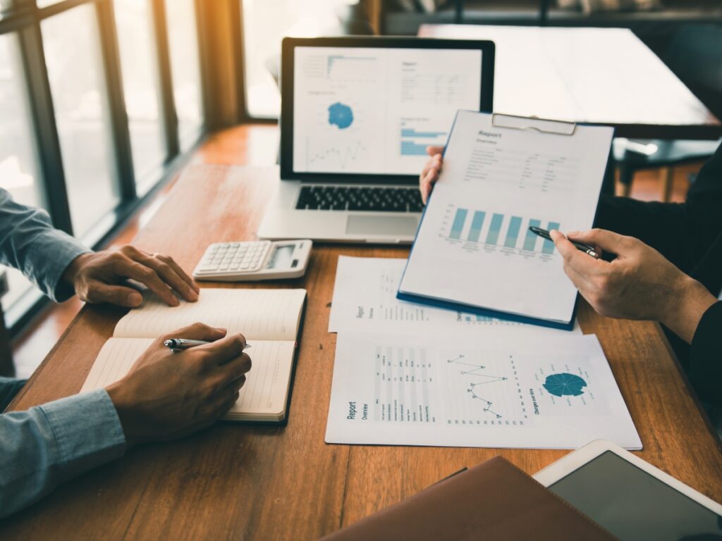 Business Fraud Investigations: How Private Investigators Help Protect Your Company 3 Two people review charts, graphs, and financial reports at a wooden table with a laptop, calculator, clipboard, and notebook in a bright office setting as part of business fraud investigations.