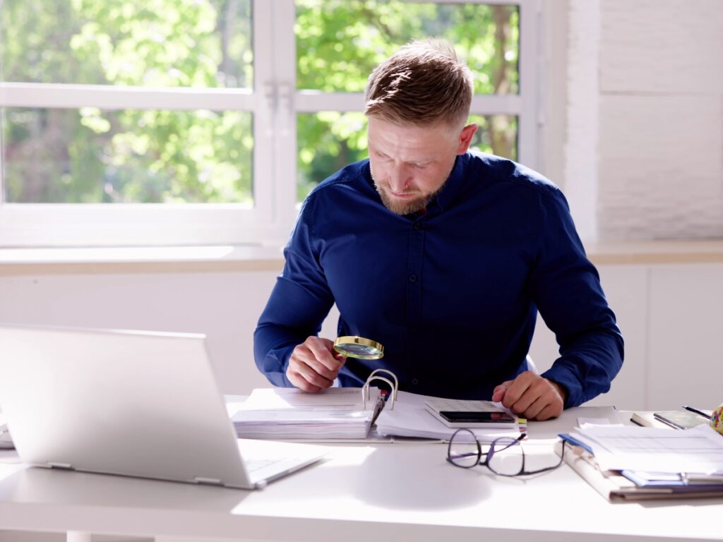 Business Fraud Investigations: How Private Investigators Help Protect Your Company 7 A man in a blue shirt examines documents with a magnifying glass at a desk, conducting business fraud investigations with a laptop, phone, and papers in front of him.